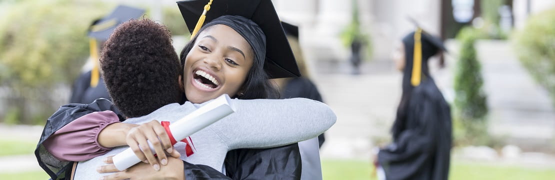 Black female college graduate hugs a loved one while holding a diploma and smiling widely.