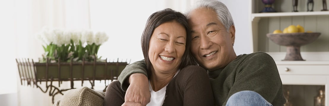 Older Asian couple embraces warmly on a living room couch.